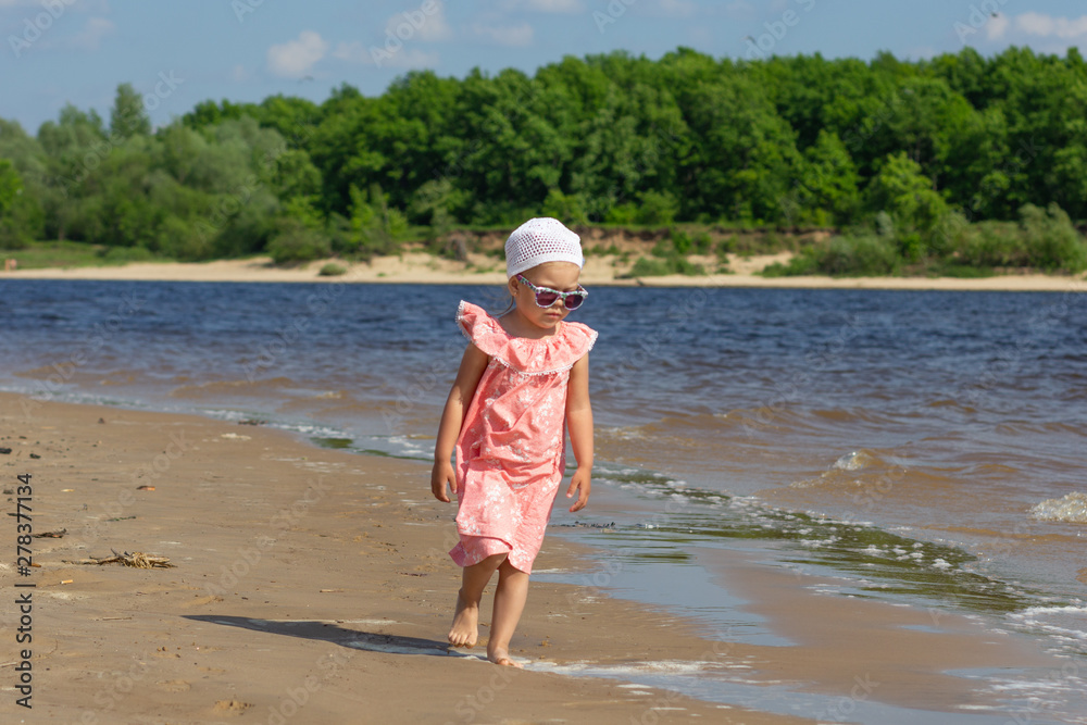 Little girl walking on the summer beach washed by waves with beautiful landscape on background Little girl walking on the summer beach washed by waves with beautiful landscape on background