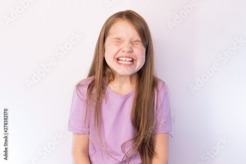 The lovely little girl, in a lilac undershirt and with a flowing hair, having strongly blinked, shows teeth, on a light background