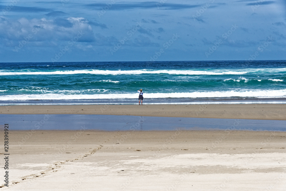 Frau blickt hinaus auf das stürmische Meer ein Fusspur im Sand führt zu ihr.