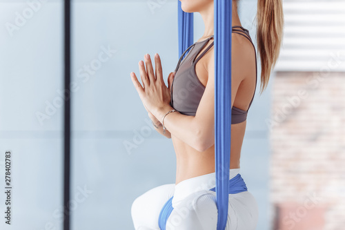 Beautiful young female fitness instructor meditates sitting in lotus pose in blue hammock in gym with large windows. Relaxation pose after yoga working out, meditating, resting, namaste gesture photo