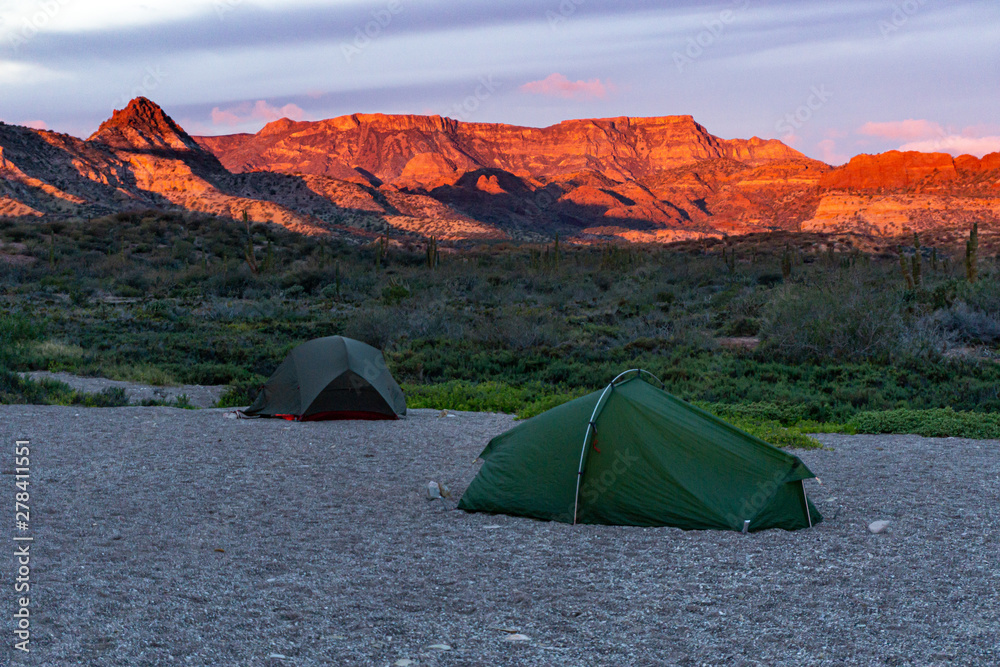 green tent in a remote valley with.a wide mountain range in the background at a dramatic .sunset