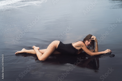 Sexy blond girl in black swimwear rests on black sand beach.