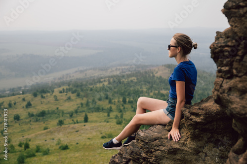 Young beautiful blonde tourist girl sits on a rocky ledge of a rock and looks far into the distance in the early foggy morning.