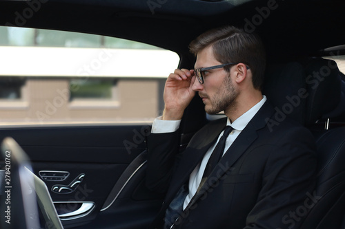 Thoughtful confident businessman keeping hand on glasses while sitting in the luxe car. © ty