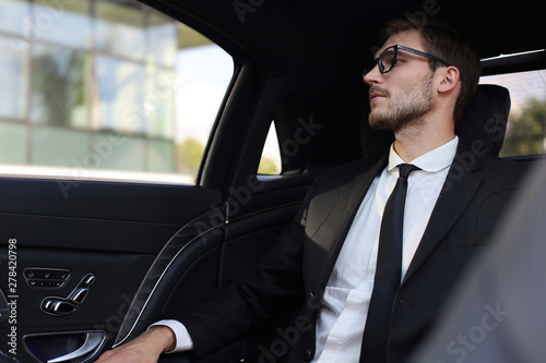 Thoughtful confident businessman in full suit with eyewear looking away while sitting in the car.