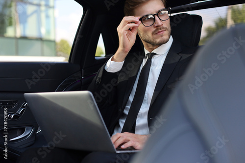 Thoughtful confident businessman keeping hand on glasses while sitting in the luxe car and using his laptop.