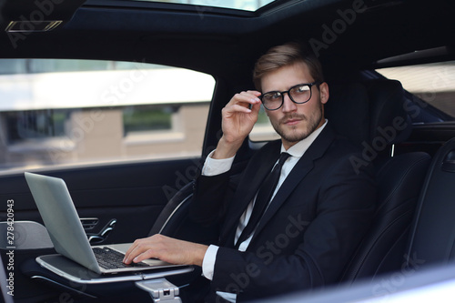 Thoughtful young businessman keeping hand on glasses while sitting in the lux car and using his laptop.