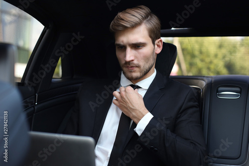 Handsome confident man in full suit straightens tie while working on laptop and sitting in the car. © ty