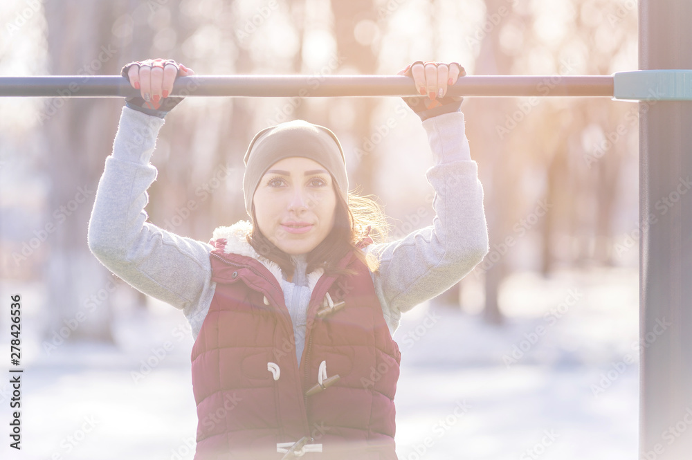 Fototapeta premium Attractive girl involved in sports workout on a horizontal bar on a sunny winter day. The concept of sports activities all year round