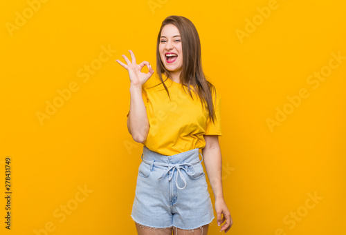 Young woman teenager wearing a yellow shirt winks an eye and holds an okay gesture with hand.