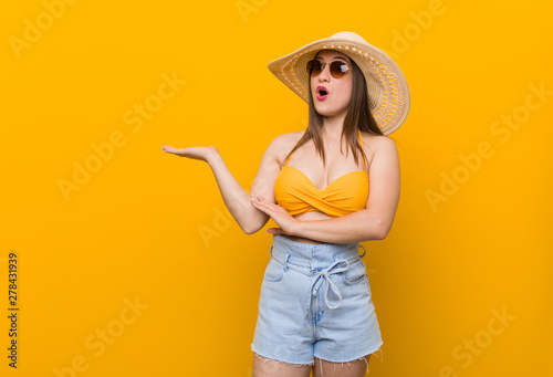 Young caucasian woman wearing a straw hat, summer look impressed holding copy space on palm.
