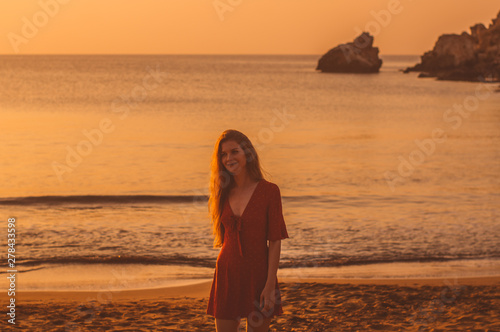 Young smiling blonde girl in red dress walking in the evening on the beach