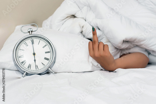 Hand under blanket showig middle finger, offensive gesture. Fu ck you concept. for alarm clock, Photo of young couple sleeping in bed while woman stretching hand to alarm clock to turn it of. photo