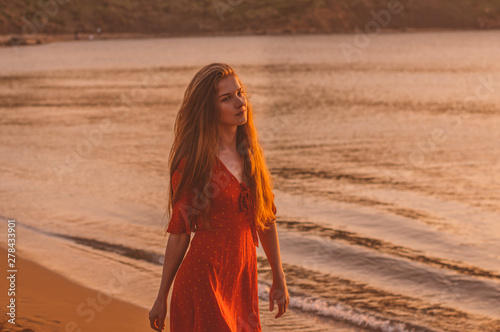 Young blonde girl in red dress walking in the evening on the beach
