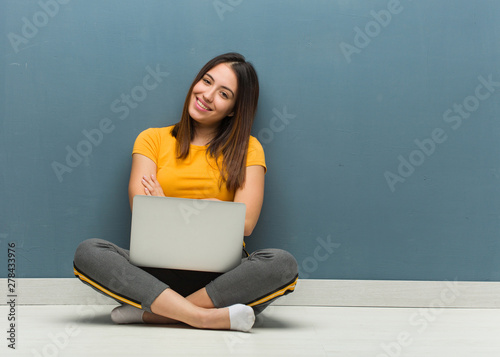 Young woman sitting on the floor with a laptop crossing arms, smiling and relaxed