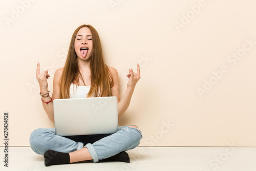 Young ginger woman sitting on her house floor showing rock gesture with fingers © Asier