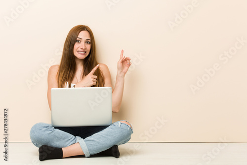 Young ginger woman sitting on her house floor shocked pointing with index fingers to a copy space. © Asier