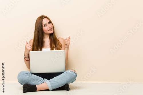 Young ginger woman sitting on her house floor raising both thumbs up, smiling and confident. © Asier