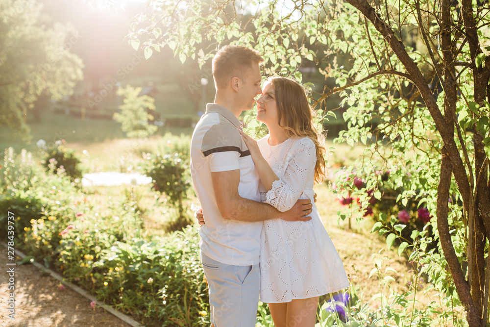 Young pretty couple in love standing in park. Handsome cheerful blonde girl in white dress hugging her boyfriend. Man and woman having fun outdoors Young pretty couple in love standing in park. Handsome cheerful blonde girl in white dress hugging her boyfriend. Man and woman having fun outdoors