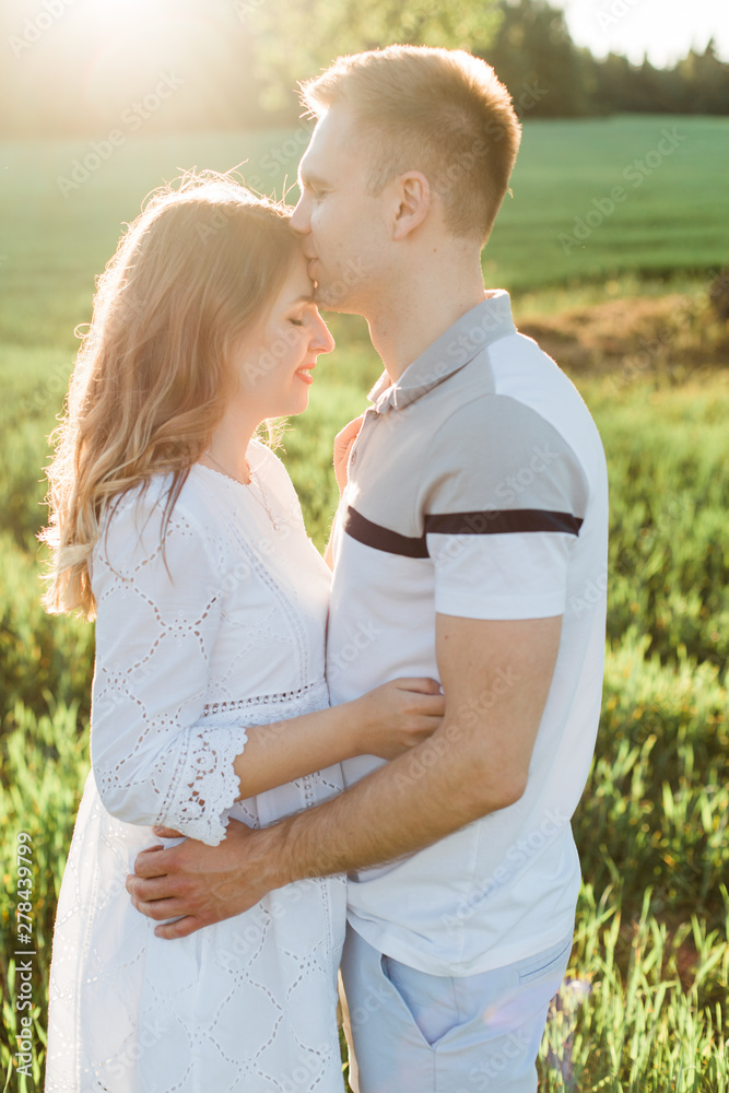 Obraz premium Young pretty couple in love standing in park. Handsome cheerful blonde girl in white dress hugging her boyfriend. Man and woman having fun outdoors
