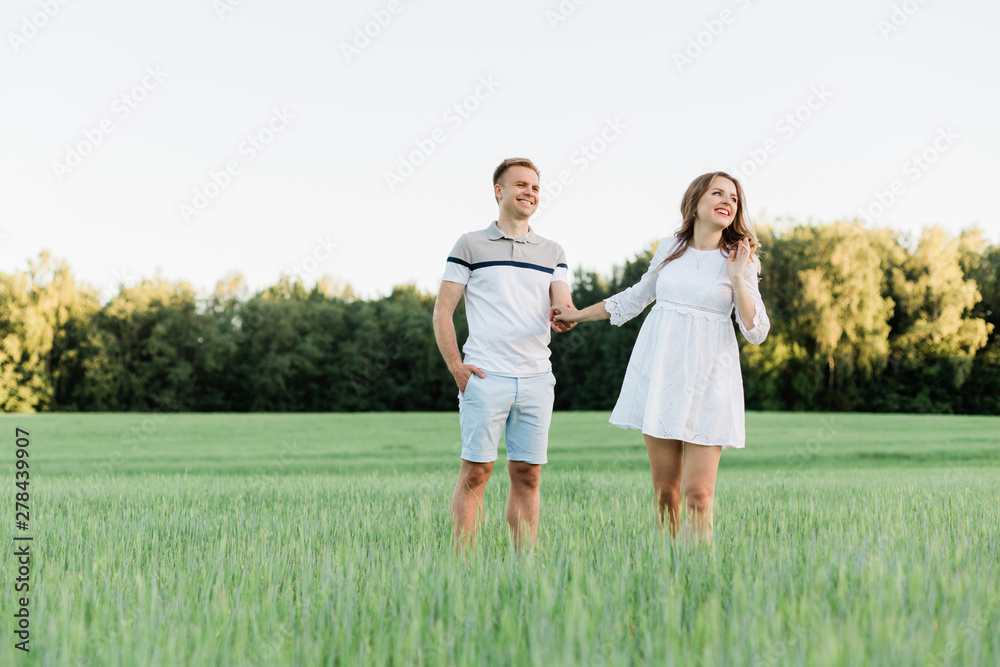 Young pretty couple in love standing in the field. Handsome cheerful blonde girl in white dress hugging her boyfriend. Man and woman having fun outdoors Young pretty couple in love standing in the field. Handsome cheerful blonde girl in white dress hugging her boyfriend. Man and woman having fun outdoors
