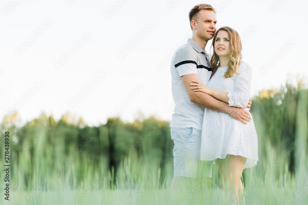 Young pretty couple in love standing in the field. Handsome cheerful blonde girl in white dress hugging her boyfriend. Man and woman having fun outdoors Young pretty couple in love standing in the field. Handsome cheerful blonde girl in white dress hugging her boyfriend. Man and woman having fun outdoors