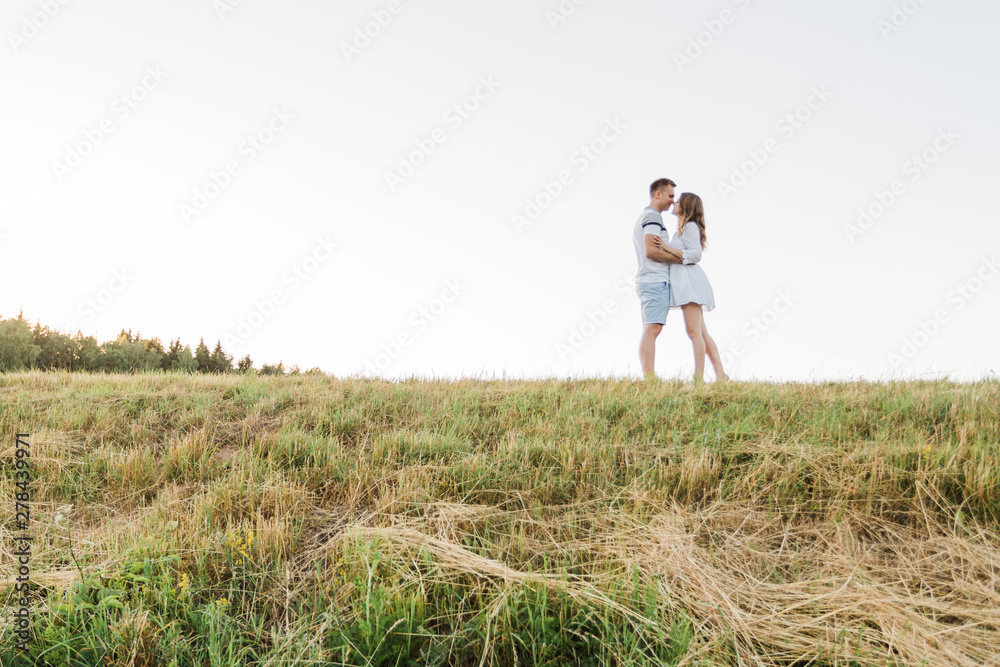 Young pretty couple in love standing in the field. Handsome cheerful blonde girl in white dress hugging her boyfriend. Man and woman having fun outdoors Young pretty couple in love standing in the field. Handsome cheerful blonde girl in white dress hugging her boyfriend. Man and woman having fun outdoors