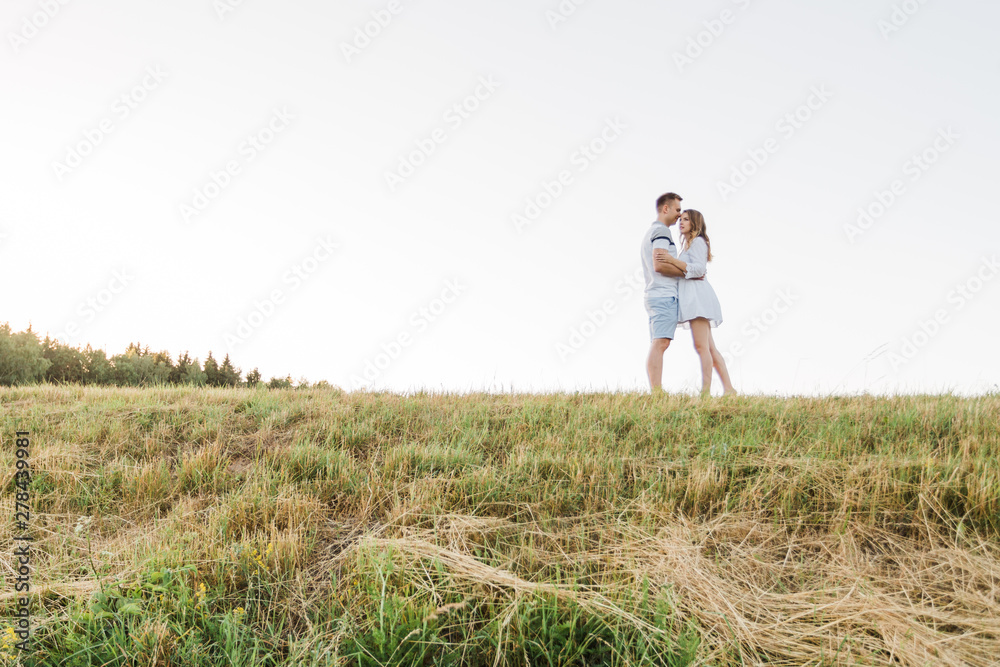 Young pretty couple in love standing in the field. Handsome cheerful blonde girl in white dress hugging her boyfriend. Man and woman having fun outdoors Young pretty couple in love standing in the field. Handsome cheerful blonde girl in white dress hugging her boyfriend. Man and woman having fun outdoors