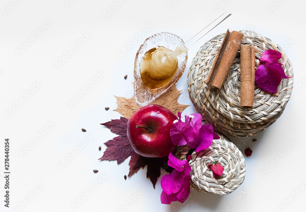 Rosh Hashanah Jewish holiday concept - red apple, glass dipper, saucer of honey on autumn maple leaves, white background with pink flowers, wicker baskets and cinnamon. Traditional holiday symbols