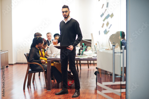 Face of handsome arabian business man, holding clipboard and glasses of beverage on the background of business peoples multiracial team meeting, sitting in office table.