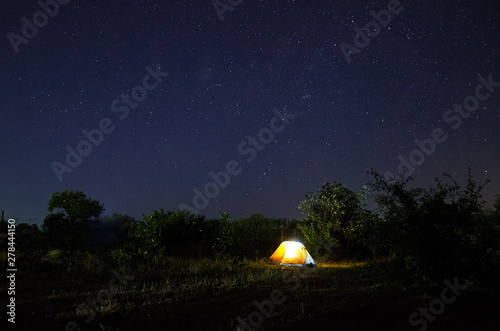 Camping tent under beautiful night sky full of stars. Starry night sky above illuminated touristic tent.