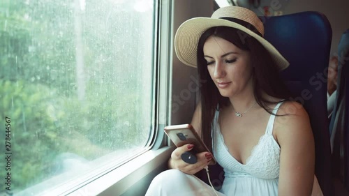Wallpaper Mural Beautiful long dark hair woman in white summer dress and straw hat sitting on train near the window and holding modern smartphone in her hands. Fast commuter vehicle riding with high speed and passing Torontodigital.ca