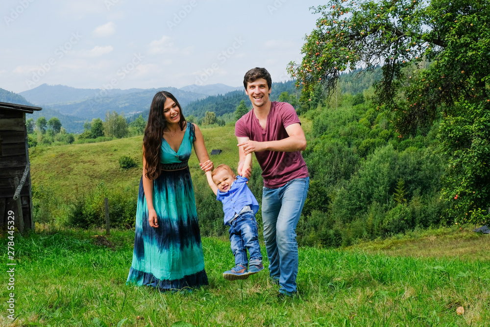 Fototapeta premium Happy young family and little son playing, holding hands and having fun outdoors. Green mountains on background. Lifestyle. Sincere moments of family. Leisure, childhood, parenthood and enjoying life