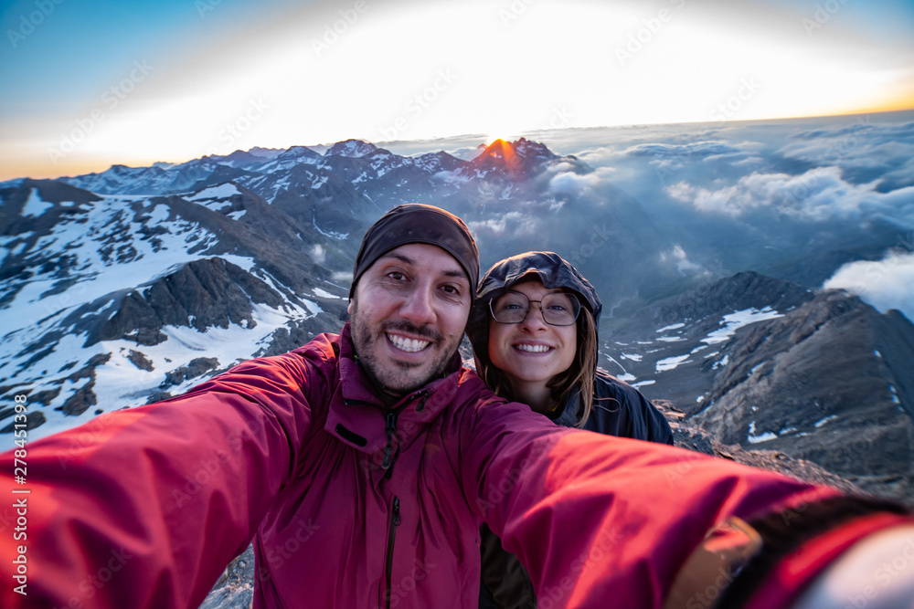 Two friends are a group of hikers having fun and making selfie on the top of the mountain with sunrise light in background. Happy faces of people who reached success and a amazing peaks landscape