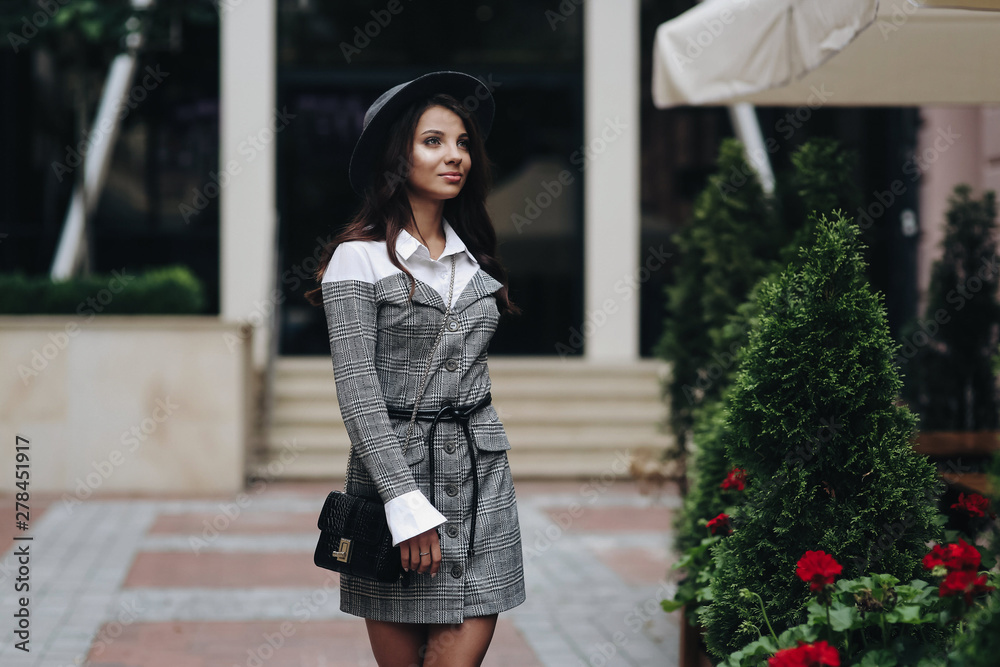 Girl outdoor posing for portrait street fashion in the urban spring city. Woman looking to the side, wearing fashionable dressed gray and white casual outfit with checkered jacket. Girl outdoor posing for portrait street fashion in the urban spring city. Woman looking to the side, wearing fashionable dressed gray and white casual outfit with checkered jacket.