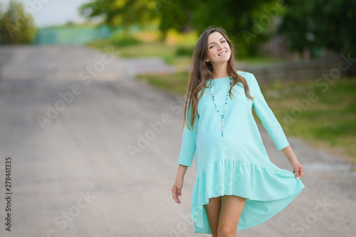Pregnant girl in a turquoise dress. Pregnant woman in day dress strolling in the park. The girl smiles. comfortable clothes for pregnant women.