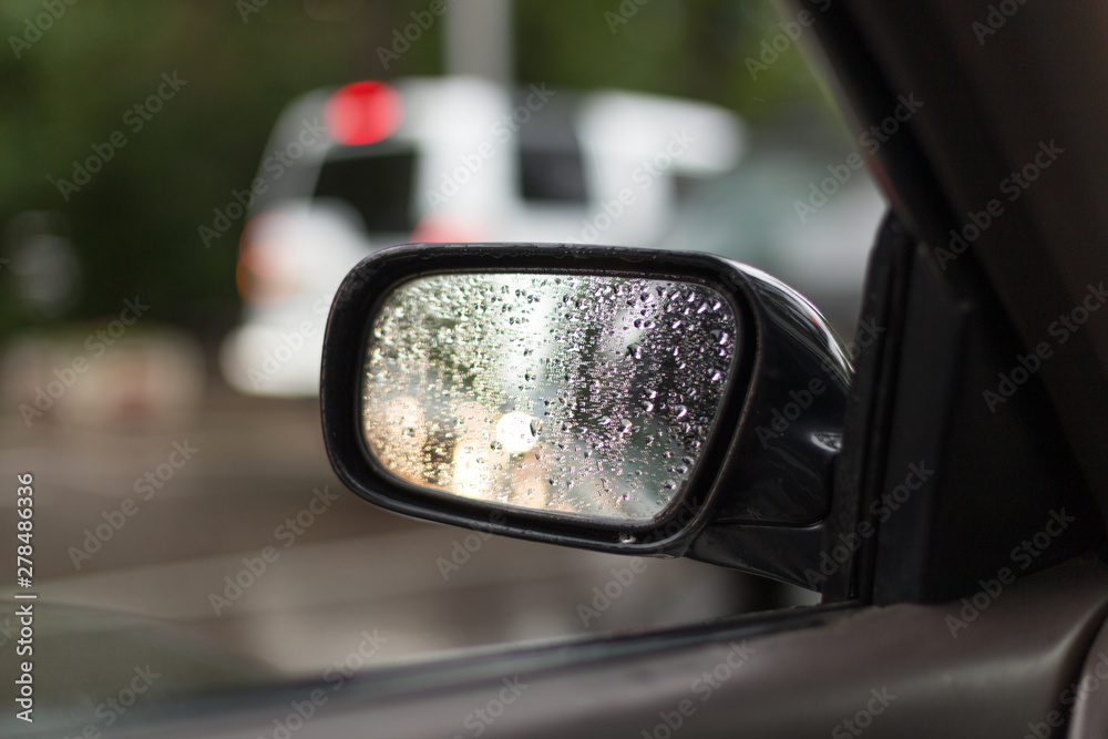 Car side mirror glass with water droplets from rain - driver's side rear view on rainy day