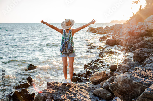 A young hipster woman in a hat and a rukzak with her hands up  standing on top of a cliff and looking at the sea at sunset.