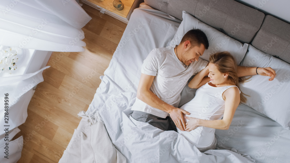 Happy Young Couple Cuddling Together in the Bed, Young Woman is Pregnant and Loving Future Father Touches and Caresses Her Belly Tenderly. Top Down Shot. Happy Young Couple Cuddling Together in the Bed, Young Woman is Pregnant and Loving Future Father Touches and Caresses Her Belly Tenderly. Top Down Shot.