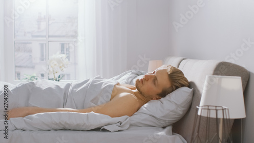 Handsome Fair Boy Lies Cozily in Bed, Opens His Eyes. Depressed Young Caucasian Man. Early Morning Sun Shines Through the Window 
