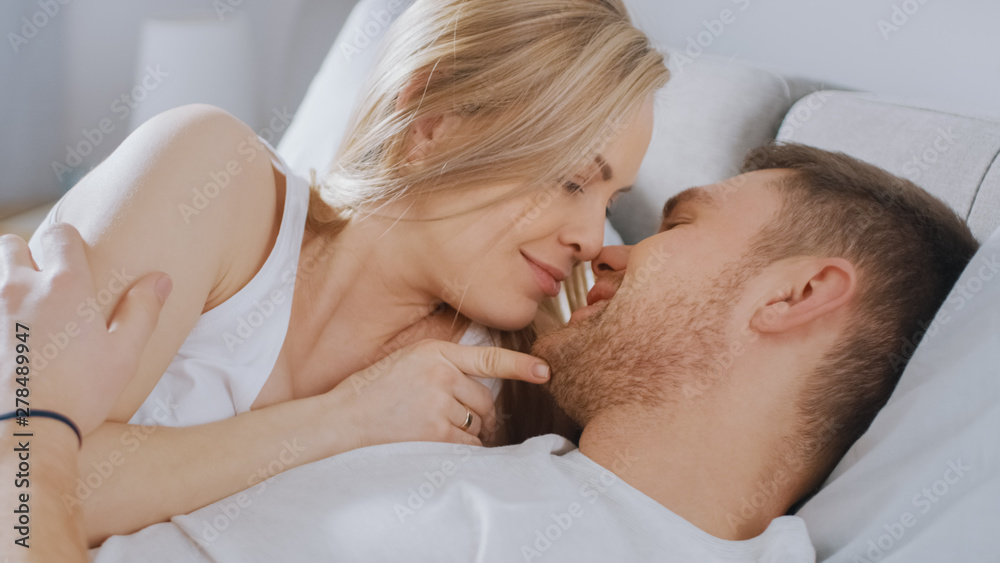 Happy Young Couple Cuddling Together in the Bed, Kissing and Smiling. Beautiful Blonde Female and Her Fit Loving Partner Experiencing Tender and Loving Moments of Living Together. Happy Young Couple Cuddling Together in the Bed, Kissing and Smiling. Beautiful Blonde Female and Her Fit Loving Partner Experiencing Tender and Loving Moments of Living Together.