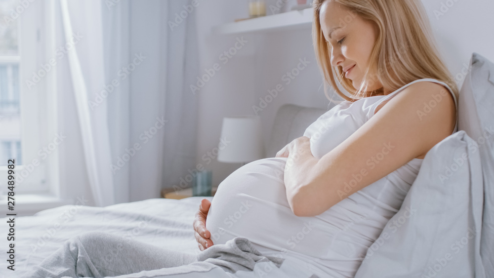 Happy Young Pregnant Woman Laying in Bed in the Morning, She Touches and Caresses Her Belly Tenderly, Loving Her Future Baby and Every Moment of Her Life. Happy Young Pregnant Woman Laying in Bed in the Morning, She Touches and Caresses Her Belly Tenderly, Loving Her Future Baby and Every Moment of Her Life.