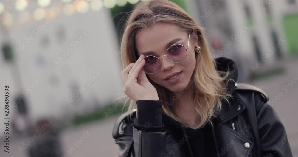 Young pretty girl smiling and posing on camera, near a modern residential area, evening in the city, blurry lights on background, positive emotions.