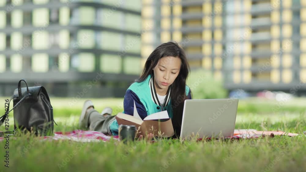Asian female college student lying on blanket on grass and doing homework on campus. Girl browsing through book and using laptop computer, another student passing by