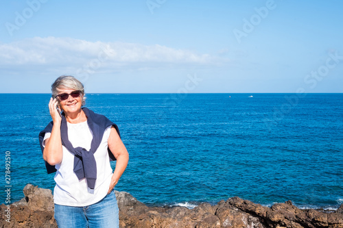 Smiling senior woman standing on the cliff over the sea. Blue sky and water. Casual clothing talking at phone. Freedom and relaxation