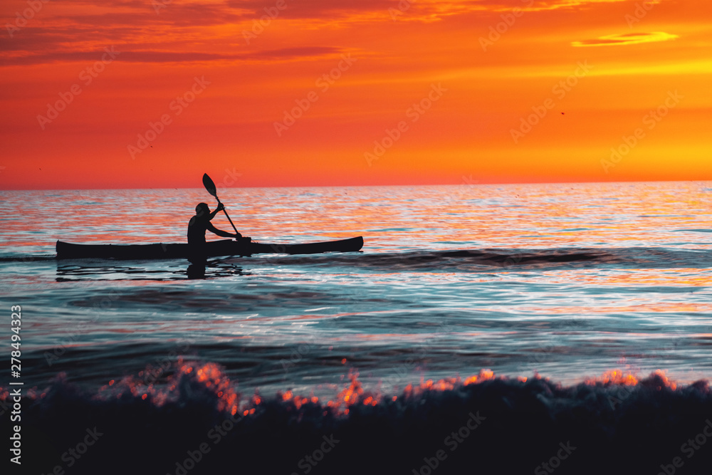 A silhouette of a man paddling in a canoe at colorful sunset light and calm ocean water. Active water sports. Rubjerg Knude Lighthouse, Lønstrup in North Jutland in Denmark, Skagerrak, North Sea