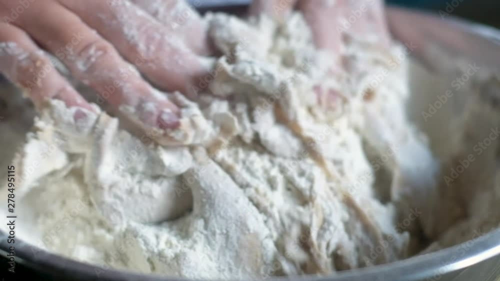 woman confectioner in an apron with symbols hands mixes soft, sticky dough in a metal bowl and sprinkled with flour. Soft evening lighting. Natural light. Panorama shooting. woman confectioner in an apron with symbols hands mixes soft, sticky dough in a metal bowl and sprinkled with flour. Soft evening lighting. Natural light. Panorama shooting.