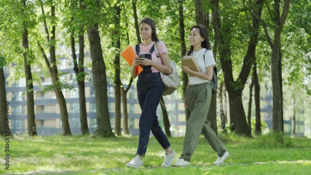 Full length tracking shot of two diverse female college students, Asian and Caucasian, with backpacks, textbooks and binders talking while walking through green park after classes Full length tracking shot of two diverse female college students, Asian and Caucasian, with backpacks, textbooks and binders talking while walking through green park after classes