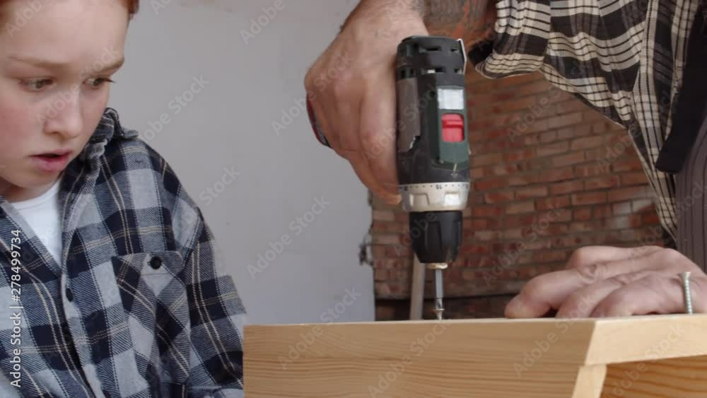 Close up of curious Caucasian red-haired girl watching attentively good-looking senior man screwing screws in wooden planks with electrical hand screwdriver Close up of curious Caucasian red-haired girl watching attentively good-looking senior man screwing screws in wooden planks with electrical hand screwdriver