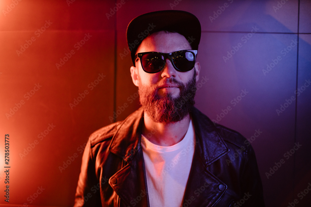 Hipster handsome man on the city streets being illuminated by neon signs. He is wearing leather biker jacket or asymmetric zip jacket with black cap, jeans and sunglasses. Hipster handsome man on the city streets being illuminated by neon signs. He is wearing leather biker jacket or asymmetric zip jacket with black cap, jeans and sunglasses.